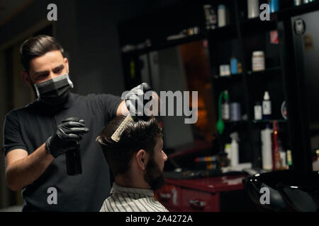 Handsome bearded man sitting in chair and covered by striped cape at barbershop while male hairdresser in black shirt looks very concentrated during working process. Male beauty treatment Stock Photo