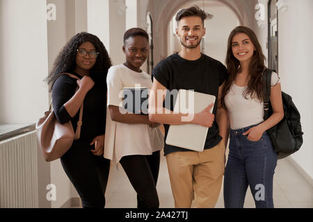 Group of four mixed-race smiling students with laptop, notebook and backpacks looking at camera in university hall. Young attractive people are gathering together to work on scientific project Stock Photo