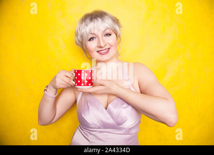 pretty curvy young blonde woman with short hair enjoys drinking tea from cute red polka dot ceramic cup on yellow studio background. Stock Photo