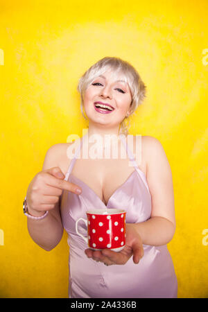 pretty curvy young blonde woman with short hair enjoys drinking tea from cute red polka dot ceramic cup on yellow studio background. Stock Photo