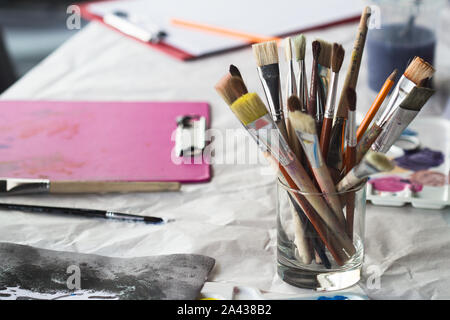 horizontal closeup perspective view of handful of brushes of different types in a glass jar on a children painting workshop white table covered with p Stock Photo