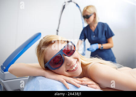 Cosmetology Service. Young woman in safety goggles at beauty clinic lying on medical bed smiling cheerful close-up while doctor preparing device Stock Photo