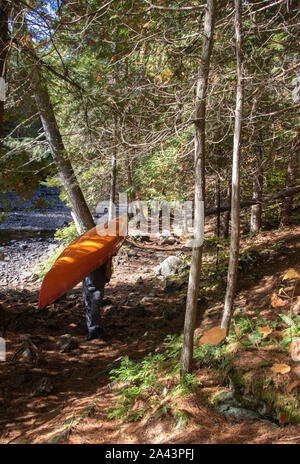 Man carrying an orange canoe along a rugged trail beside a river Stock Photo