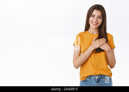 Tender, charming caucasian woman in yellow t-shirt, holding hands pressed to heart, cherish relationship and love, smiling joyfully, dreamy look camer Stock Photo