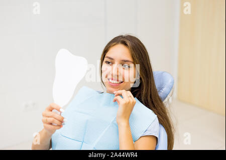 The patient admires her smile looking at the mirror sitting in dental chair after dental check. Stock Photo