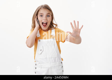 Amused cute excited caucasian little girl, child talking on smartphone, use mobile phone talking best friend with playful happy grin, raise hand show Stock Photo