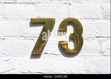 Golde colored house number seventy three (73) on a white painted brick wall. Stock Photo