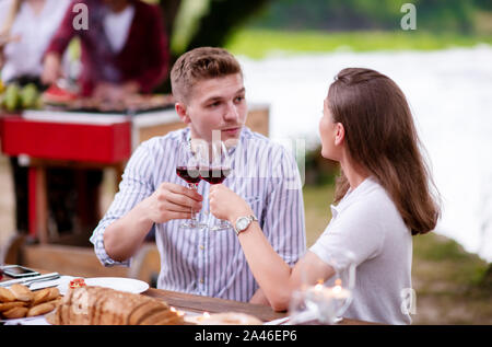 young happy couple toasting red wine glass while having picnic french dinner party outdoor during summer holiday vacation  near the river at beautiful Stock Photo