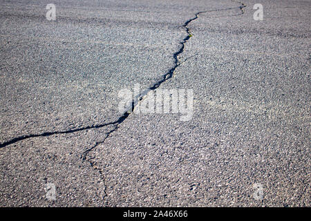 A road paved with asphalt has a large crack across it, which forks into two in the foreground. Stock Photo