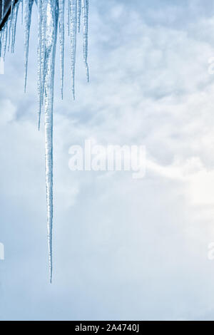 Close-up of group of transparent icicles hanging from the roof. One of them is unusually large, thick and sharp. Beautiful cloudy sky at background. W Stock Photo