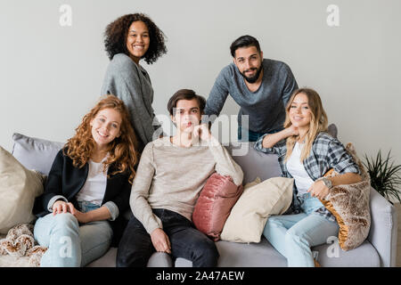 Group of happy young intercultural friends in casualwear relaxing on soft couch Stock Photo
