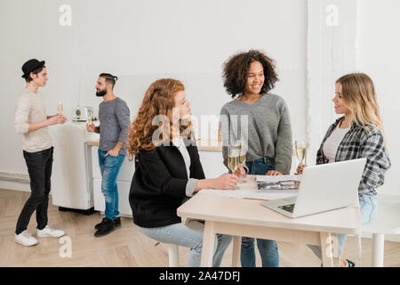 Three young casual women with champagne discussing something by desk Stock Photo