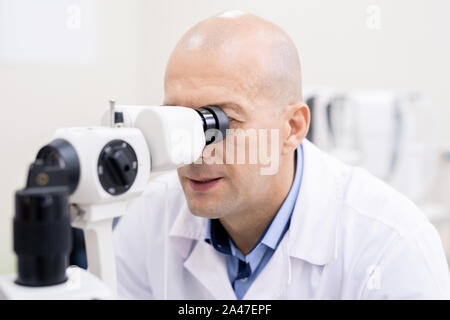 Professional ophthalmologist looking through medical equiment by workplace Stock Photo