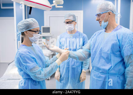 Young nurse helping professor in protective uniform to put on gloves in hospital Stock Photo