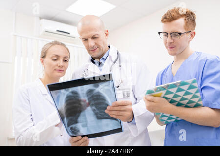 Two doctors discussing x-ray of lungs while young intern making notes Stock Photo