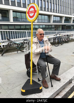 Elderly male / senior man waiting patiently on his portable push frame seat for his bus to arrive at his temporary bus stop on the pavement. Twickenham. UK. (stockmo) Stock Photo