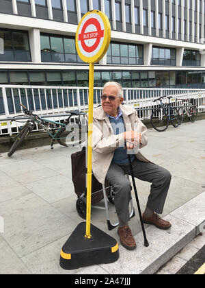 Elderly male / senior man waiting patiently on his portable push frame seat for his bus to arrive at his temporary bus stop on the pavement. Twickenham. UK. (stockmo) Stock Photo