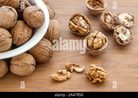 Natural unbleached walnuts (Juglans regia) in a white bowl, cracked nuts and kernels on a brown wooden board. Vegetarian food. Healthy eating. Stock Photo