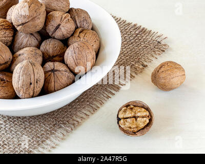 Natural unbleached walnuts (Juglans regia) in a white bowl on a rustic style burlap table napkin over withe rough background. Vegetarian food. Stock Photo
