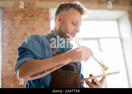 Serious mature painter in workwear mixing colors with paintbrush on palette Stock Photo