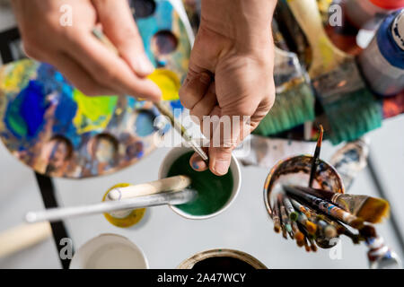 Hands of painter holding paintbrush over glass of water while cleaning it Stock Photo