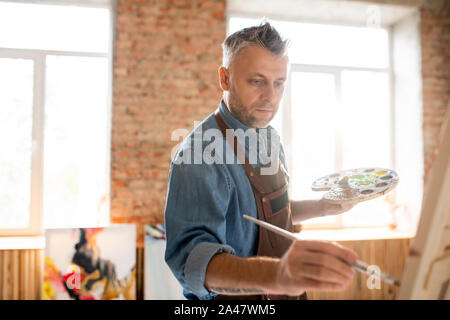 Inspired man with palette and paintbrush standing in front of easel and painting Stock Photo