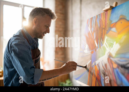 Happy mature master of arts in apron working over abstract painting Happy mature master of arts in apron working over abstract painting