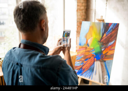 Creative man standing in front of painting while taking its photo on smartphone Stock Photo