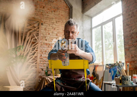 Contemporary mid-aged artist sitting on chair in studio and choosing paintbrush Stock Photo