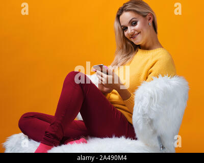 Happy and cheerful blonde uses smartphone for communication and online shopping. Positive young woman in a yellow sweater on a yellow background. Stock Photo