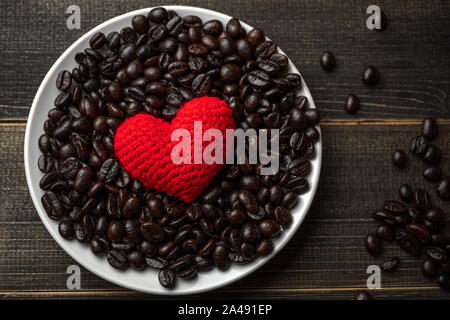 red heart on a lot of roasted coffee beans background on white plate on wooden table. Strong black espresso, Grains of coffee background, texture Stock Photo