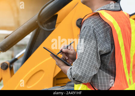Engineering wearing a white safety helmet standing In front of the backhoe And are using tablet for check the blueprint with construction Stock Photo