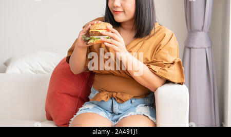 Hungry overweight woman smiling and holding hamburger and sitting in the living room, her very happy and enjoy to eat fast food. Concept of binge eati Stock Photo