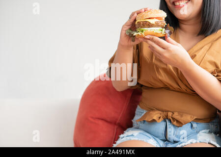 Hungry overweight woman smiling and holding hamburger and sitting in the living room, her very happy and enjoy to eat fast food. Concept of binge eati Stock Photo
