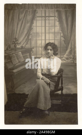 Edwardian studio portrait, cut down postcard of pretty young woman or late teens girl seated by piano prop, wearing high necked lace blouse and woollen skirt, circa 1910, 1911, U.K. Stock Photo