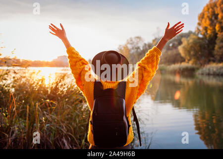 Traveler with backpack relaxing by autumn river at sunset. Young woman raised arms feeling free and happy Stock Photo