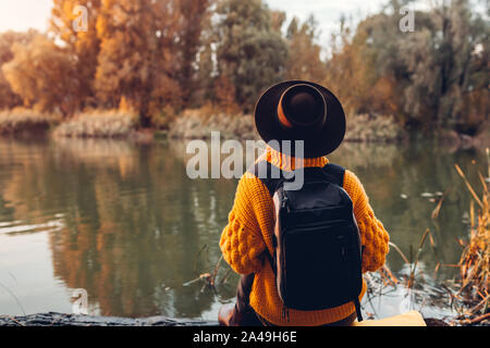 Traveler with backpack relaxing by autumn river at sunset. Young woman sitting on bank and relaxing Stock Photo