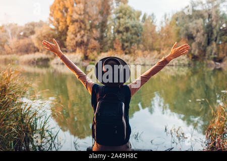 Traveler with backpack relaxing by autumn river at sunset. Young woman raised arms feeling free and happy Stock Photo