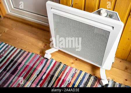 White electric radiator with a thermostat is placed on the floor covered with a colored rug, next to a wall and door, in a wooden country house. Stock Photo