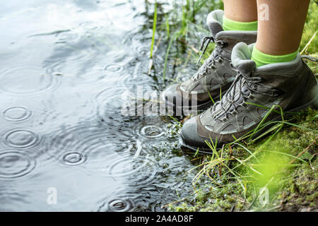Woman in tourist waterproof hiking boots is standing on the shore of a lake, right next to the water in the rain. Close-up. Stock Photo