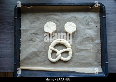 Two pieces of raw dough and a pretzel are laid out on a tray in the form of a sad face, against a textured wooden table. Top view. Stock Photo