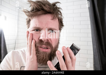 A man holds a trimmer in his hands looking at his reflection in the mirror and is horrified by his shaggy hairstyle and beard, early in the morning, a Stock Photo