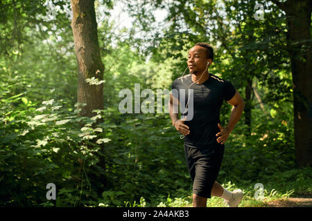 African man with sportive body running in garden in summer morning. Sportsman wearing in black t shirt and shorts listening music with headphones. Runner training. Stock Photo