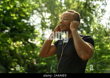 Close up of african sportsman listening music with headphones, enjoying. Muscular runner running in forest with closed eyes. Man with sportive body wearing in black t shirt.