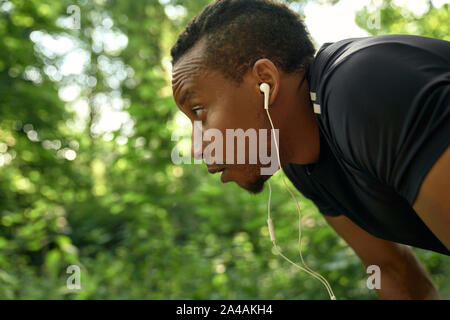 Close up of muscular runner with curly hair wearing in black t shirt outdoors. Handsome african sportsman listening music with headphones. Athlete posing side view, looking forward.
