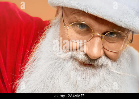 Close up of aged Santa Claus with real grey beard, blue eyes and wrinkles on his face looking at camera in studio. Happy grandfather in eyeglasses excited about christmas time. Stock Photo