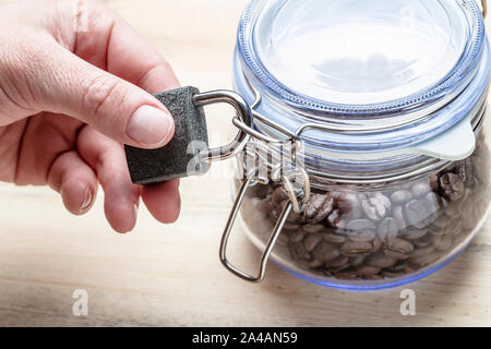 Hand is holding a metal closed lock on a transparent glass jar with a lid containing coffee beans on a wooden table background. The concept of a healt Stock Photo