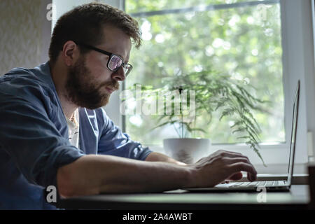 Concentrated, young, bearded man in glasses works with a laptop at home, against the window and flower, early in the morning. Stock Photo