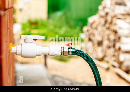 A rubber hose with a vertil for watering vegetation with water comes out of the wall of a wooden house, on a blurred fence and firewood. Close-up. Stock Photo