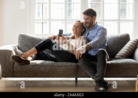 Happy couple in love relaxing on couch together, using cellphone Stock Photo